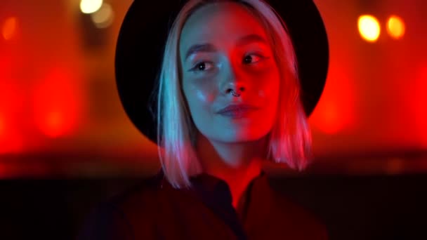 Mujer hipster con peinado rubio de pie sobre fondo brillante de neón. Ciudad por la noche. Sombrero, piercing en la nariz. Hermosa chica atractiva. Movimiento lento . — Vídeos de Stock