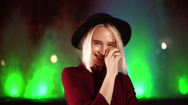 Portrét okouzlující charismatické hipster žena s blond účes stojí na pozadí neonové fontány a usmívá se. Město v noci. Klobouk, piercing v nose. Krásná atraktivní dívka. Zpomalený pohyb. — Stock video
