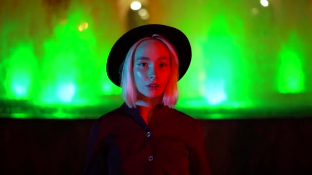 Portrait of millennial hipster woman with blond hairstyle standing on neon fountain background. City at night. Hat, nose piercing. Beautiful attractive girl. Slow motion. — Stock Video