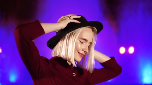 Portrait of charming charismatic hipster woman with blond hairstyle standing on neon fountain background and smiling. City at night. Hat, nose piercing. Beautiful attractive girl. Slow motion. — Stock Video