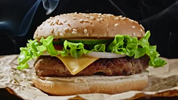 Μεγάλο ορεκτικό burger με κοτολέτες κρέατος, κρεμμύδι, λαχανικά, λιωμένο τυρί, μαρούλι και σάλτσα μαγιονέζας σε περγαμηνή. Μεμονωμένο χάμπουργκερ περιστρέφεται σε σκούρο φόντο καπνού, κοντινή προβολή — Αρχείο Βίντεο