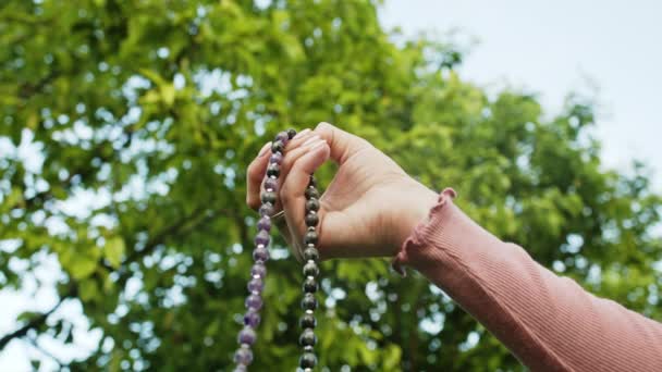 Hand einer jungen gläubigen Frau betet in der Natur und verwendet handgefertigte Rosenkranzperlen, um Gebete zu zählen und sich auf die Meditation zu konzentrieren. Religion, Hoffnung, Liebeskonzept. Zeitlupe. — Stockvideo