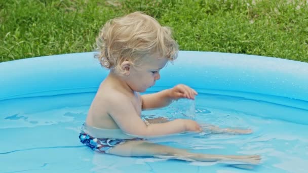 Carino bambino che fa il bagno nella piscina blu della strada nel cortile. Ritratto di bambino gioioso, baby. Il ragazzo ride, spruzza acqua, sorride. Concetto di stile di vita sano, famiglia, tempo libero in estate. Rallentatore. — Video Stock