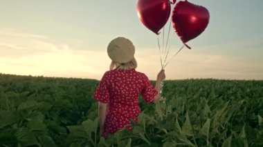 Yeşil alanda kalp şeklinde balonlarla poz veren kırmızı elbiseli güzel bir kadın. Hasır şapkalı ya da bereli bir kız. Doğum günü, tatil, özgürlük kavramını kutlamak.