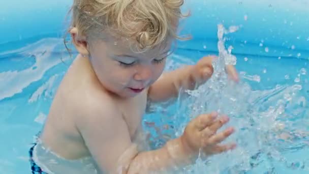 Милый маленький ребенок купается в бассейне на голубой улице во дворе. Портрет радостного малыша, детка. Малыш смеется, брызгает водой, улыбается. Концепция здорового образа жизни, семьи, досуга летом. Медленное движение . — стоковое видео