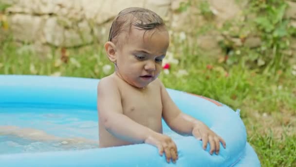 Очаровательный маленький ребенок плавает в бассейне на голубой улице во дворе. Портрет радостного малыша. Малыш смеется, брызгает водой, улыбается. Концепция здорового образа жизни, семьи, досуга летом. Медленное движение . — стоковое видео