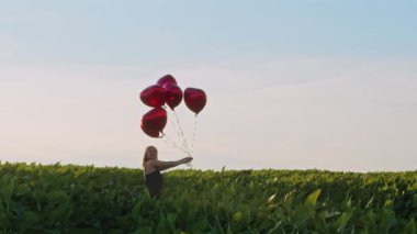 Yeşil alanda kalp şeklinde balonlarla poz veren kırmızı elbiseli güzel bir kadın. Hasır şapkalı ya da bereli bir kız. Doğum günü, tatil, özgürlük kavramını kutlamak.