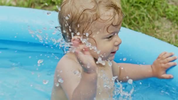 Sød lille baby svømmer i blå gade pool i gården. Portræt af glædelig lille barn. Knægten griner, sprøjter vand, smiler. Begrebet sund livsstil, familie, fritid om sommeren. Langsom bevægelse. – Stock-video