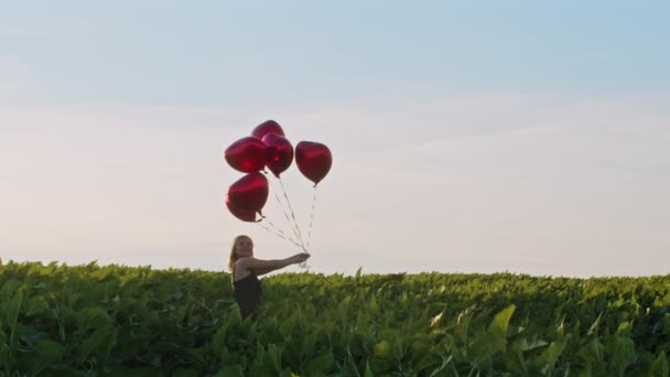 Bella donna in abito rosso retrò in posa con palloncini a forma di cuore in campo verde. Ragazza con cappello vintage di paglia o berretto. Compleanno, vacanza, celebrare il concetto di libertà. — Video Stock