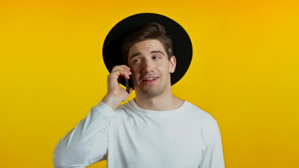 Ung man i vitt slitage och hatt talar på mobiltelefon på gul bakgrund. Den trendiga hipsterkillen har ett samtal. Smartphone, teknik koncept. — Stockvideo