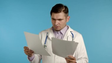 Profesyonel medikal beyaz önlüklü kafası karışmış bir adam mavi arka planda izole edilmiş dosyaları tutuyor. Sakallı ve steteskoplu bir doktor. Teşhislere nasıl tepki vereceğini bilmiyor.