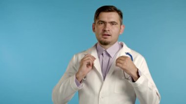 Profesyonel tıp önlüklü yakışıklı doktor portresi. Giysilerini düzeltiyor, saçlarını düzeltiyor, kameraya göz kırpıyor. Doc mavi stüdyo arka planında izole.