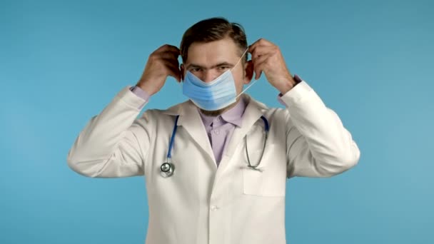 Jonge arts zet gezicht medische masker tijdens coronavirus pandemie. Medisch portret op blauwe achtergrond. Bescherming met masker tegen COVID-19-uitbraak — Stockvideo