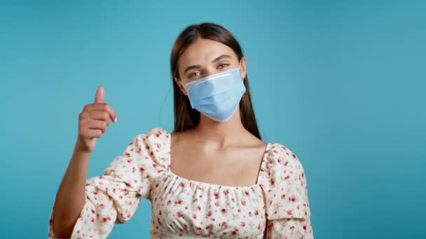 Mujer atractiva en respirador protector mostrando el pulgar hacia arriba signo sobre fondo azul. Chica joven positiva con máscara sonríe a la cámara durante la cuarentena. Ganador. Éxito. Lenguaje corporal. — Vídeos de Stock