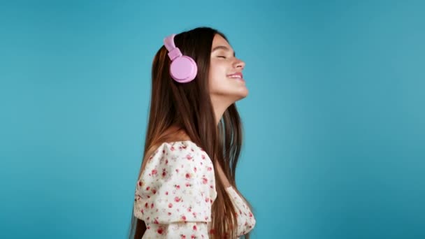 Linda chica joven con el pelo largo que se divierte, sonriendo, bailando media cara con auriculares en el estudio sobre fondo azul. Música, danza, concepto de radio, cámara lenta — Vídeo de stock