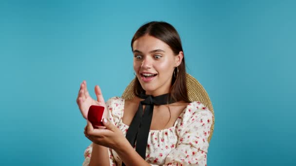 Jolie jeune femme européenne en robe tenant petite boîte à bijoux avec proposition bague en diamant sur fond de mur bleu. Fille souriante, elle est heureuse d'être présente, proposition de mariage. — Video