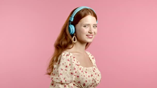 Menina bonita com cabelo vermelho se divertindo, sorrindo, dançando com fones de ouvido azuis em estúdio contra fundo rosa. Música, dança, conceito de rádio, câmera lenta — Vídeo de Stock