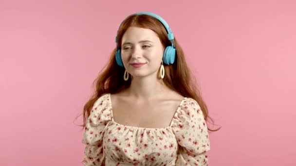 Menina bonita com cabelo vermelho se divertindo, sorrindo, dançando com fones de ouvido azuis em estúdio contra fundo rosa. Música, dança, conceito de rádio, câmera lenta — Vídeo de Stock