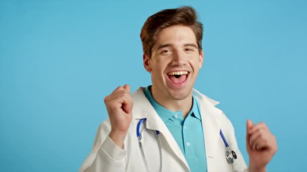 Glimlachende dokter, grappig dansen, succes en geluk op het werk. Jonge knappe dokter man in professionele medische witte jas is geïsoleerd op blauwe studio achtergrond. — Stockvideo