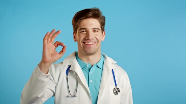 Красивый молодой человек в белом медицинском халате улыбается в камеру. Доктор показывает знак ОК на синем фоне. Победитель. Успех. Язык тела. — стоковое видео