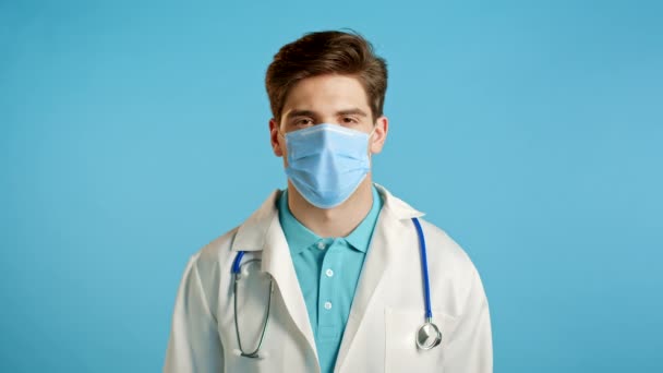 頭のジェスチャーを拒否することを示すプロの医療用マスクと白のコートで深刻な医師の肖像画、不承認記号。否定する、同意しない。青色の背景に隔離されたドク. — ストック動画