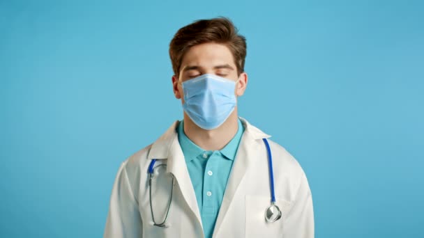 Πορτρέτο του ικανοποιημένου γιατρού σε επαγγελματική ιατρική μάσκα και παλτό δείχνει ναι σημάδι από το κεφάλι. Ο άνθρωπος το επιτρέπει, αντιμετωπίζει με επιδοκιμασία. Γιατρός απομονωμένος σε μπλε φόντο.. — Αρχείο Βίντεο