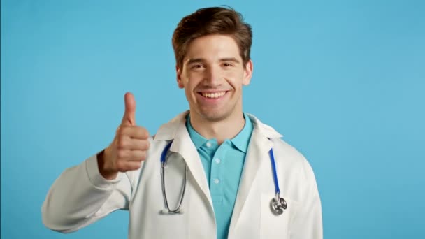 青色の背景の上に親指を示す医者。医療用コートの前向きな青年がカメラに微笑む。勝者だ。成功だ。ボディランゲージ. — ストック動画