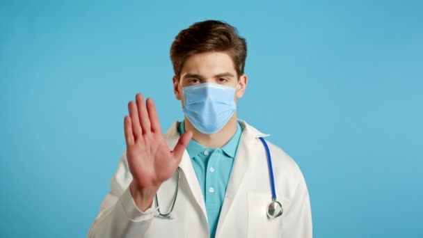 Portrait of serious doctor in professional medical white coat showing rejecting gesture by stop palm sign. Doc man isolated on blue background. — Stock Video