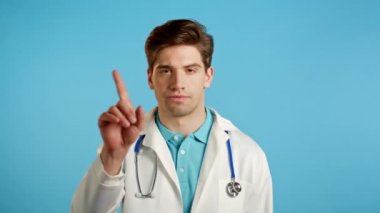 Profesyonel medikal beyaz önlüklü ciddi bir doktorun portresi reddedici bir hareket sergiliyor. Hem de parmak izi olmadan. Reddediyor, itiraz ediyor. Doktor adam mavi arkaplanda izole edilmiş..