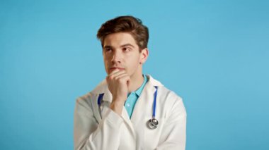 Medikal ceketli düşünceli doktor portresi mavi stüdyo arka planında bir fikir belirtiyor. Gülümseyen mutlu doktor Eureka hareketi gösteriyor..