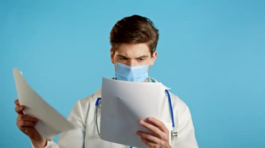 Profesyonel medikal beyaz önlüklü kafası karışmış bir adam mavi arka planda izole edilmiş dosyaları tutuyor. Maskeli ve steteskoplu bir doktor. Teşhislere nasıl tepki vereceğini bilmiyor.