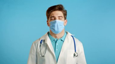 Maskeli sakin doktor dinleniyor, meditasyon yapıyor. Adam sakinleşir, derin derin nefes alır mavi stüdyo arka planında Mudra om.