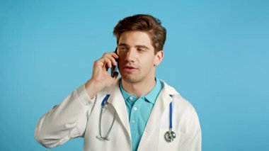 Beyaz ceketli genç doktor mavi arka planda cep telefonuyla konuşuyor. Doktor konuşacak. Akıllı telefon, teknoloji konsepti.