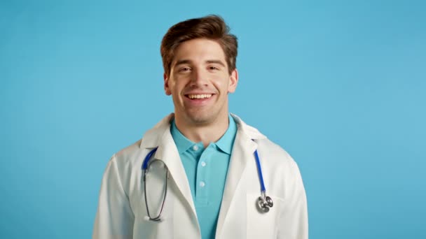 Hombre guapo doctor en ropa médica sonriendo en el fondo azul del estudio. Chico alegre riendo y mirando a la cámara. — Vídeos de Stock