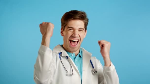 Ναι, νικήτρια χειρονομία. Ο εκπληκτικός γιατρός σοκαρίστηκε. Όμορφος γιατρός σε λευκό ιατρικό παλτό έκπληκτος στην κάμερα πάνω από το μπλε φόντο — Αρχείο Βίντεο
