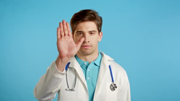 Портрет серьезного врача в профессиональном медицинском белом халате, показывающий отклоняющий жест стоп-ладонью. Доктор человек изолирован на синем фоне. — стоковое видео