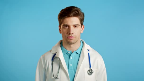 Hombre guapo doctor en bata blanca profesional sobre fondo de estudio azul. Doctor serio mirando a la cámara. — Vídeos de Stock