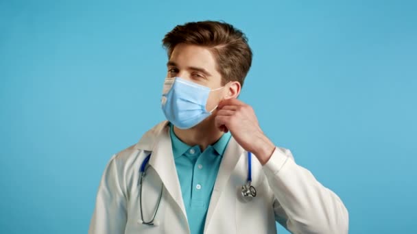Довольный врач в медицинском халате снимает защитную маску и бросает ее на пол как символ пандемии, эпидемии конца. Синий студийный фон. — стоковое видео