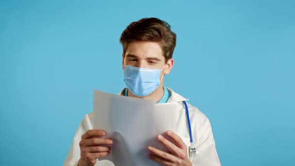 Ικανοποιημένος άνθρωπος με επαγγελματική ιατρική ποδιά και μάσκα κρατώντας αρχεία που είναι απομονωμένα σε μπλε φόντο. Κουνάει το κεφάλι του εγκρίνοντας τη διάγνωση. Γιατρός με γένια και στηθοσκόπιο.. — Αρχείο Βίντεο