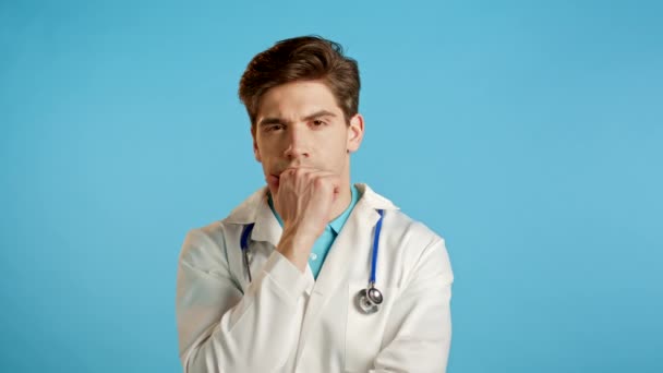 Hombre médico pensante mirando hacia arriba y alrededor sobre fondo azul. Expresiones faciales contemplativas preocupadas. Guapo doctor en abrigo médico con estetoscopio — Vídeo de stock