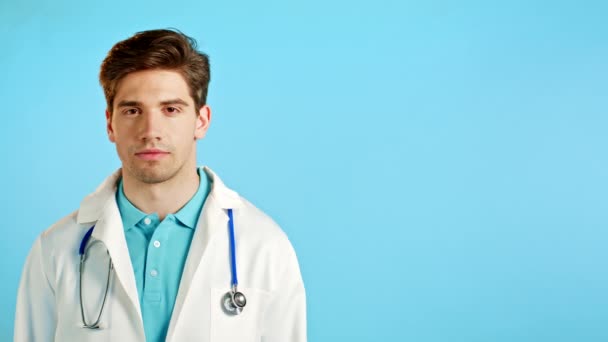 Schöner Arzt in professionellem weißen Mantel auf blauem Studiohintergrund. Seriöser Doktor blickt in die Kamera. Kopierraum. — Stockvideo