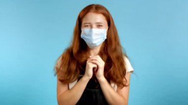 Yüzünde koruyucu maske olan kız çok mutlu ve mutlu, harika haberler sayesinde el çırpıyor. Mavi arka planda heyecanlı, mutlu bir kadın