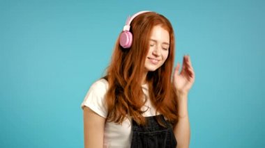 Kızıl saçlı güzel bir kız müzik dinliyor, gülümsüyor, stüdyoda pembe kulaklıklarla mavi arka planda dans ediyor. Müzik, dans, radyo konsepti, yavaş çekim.