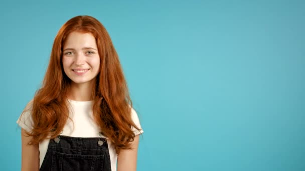 Jovem com penteado vermelho no geral olhando para a câmera. Espaço para cópia. Retrato de menina bonita bonito na parede azul estúdio fundo. — Vídeo de Stock