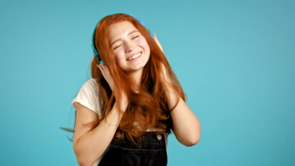 Menina com cabelos vermelhos ouvindo música, dançando energeticamente e ativamente em fones de ouvido em estúdio em fundo azul. Música, dança, conceito de rádio, câmera lenta — Vídeo de Stock