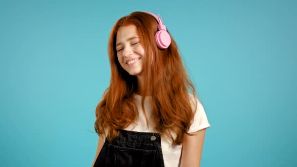 Όμορφη νεαρή κοπέλα με κόκκινα μαλλιά ακούγοντας μουσική, χαμογελώντας, χορεύοντας με ροζ ακουστικά στο στούντιο με μπλε φόντο. Μουσική, χορός, ραδιοφωνική ιδέα, αργή κίνηση. — Αρχείο Βίντεο