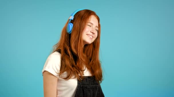 Bella ragazza dai capelli rossi che ascolta musica, sorride, balla in cuffie blu in studio sullo sfondo. Musica, danza, concetto radio, slow motion. — Video Stock