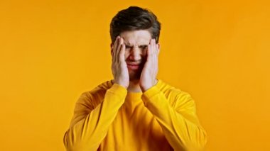 Baş ağrısı çeken genç bir adam, stüdyo portresi. Adam ellerini başının üstüne koyuyor, sarı arka planda izole edilmiş. Sorunlar, ilaç, hastalık kavramı