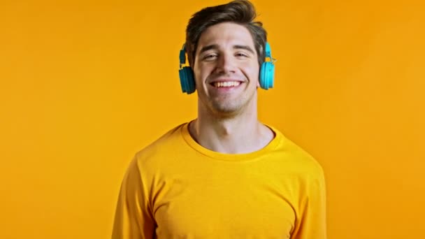 Hezký mladý muž s módním účesem baví, usmívá se, tančí s modrými sluchátky ve studiu na žlutém pozadí. Hudba, tanec, koncept rádia, zpomalení — Stock video