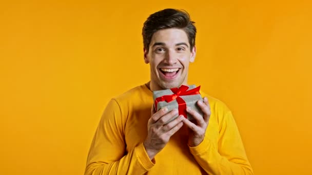 Hombre guapo sosteniendo caja de regalo en el fondo amarillo del estudio y sonríe a la cámara. feliz europeo hipster chico recibido presente e interesado en lo que es dentro de. — Vídeos de Stock